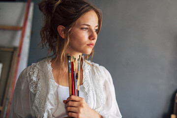 Wall Mural - Portrait of an artist female standing next to the easel with canvas painting in her art studio. A young woman student painter searching for an imagination at home.