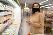 © Andrii Lysenko - Close-up portrait, a young lady in a supermarket stands near a shop window, a protective mask is put on her face. Pandemic shopping concept.