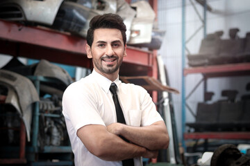 Wall Mural - Portrait of smiling businessman seller standing with arms crossed at auto spare parts store shop warehouse with many second hand engine parts as blurred background.