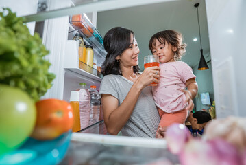 asian mother carrying her daughter while taking a glass of juice inside the fridge