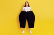 Shocked astonished woman wearing old too big black trousers keeps hands in pants. looks at camera with open mouth and big eyes, has surprised facial expression, posing isolated over yellow background.