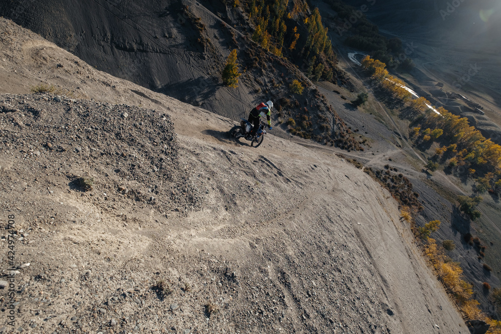 Extreme Rider climbing sand mountain top on off-road cross enduro motorcycle. Beautiful mountains landscape down on background, colourful autumn forest and river in sunshine - obrazy, fototapety, plakaty 