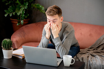 Wall Mural - Feeling tired. Frustrated young  man looking exhausted and covering his face with hands while sitting at laptop his home working place