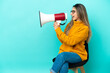 © luismolinero - Young caucasian woman sitting on a chair isolated on blue background shouting through a megaphone