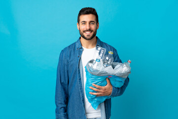 Young caucasian man holding a bag full of plastic bottles to recycle isolated on blue background smiling a lot