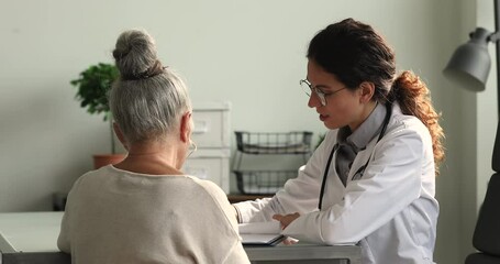 Wall Mural - Back rear view focused senior grandmother visiting professional female 35s general practitioner in clinic office, consulting about disease treatment or medical insurance contract, healthcare concept.