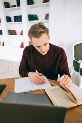 Young caucasian man college student studying with laptop distantly preparing for test exam writing essay doing homework at home or college library.