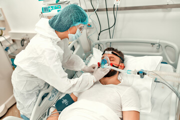 Doctors in protective suits put on a ventilation mask on a sick man with coronavirus disease covid-19, who is in an intensive care unit in a modern hospital.