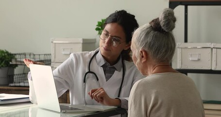 Wall Mural - Professional young 35s female physician showing medical insurance presentation or health test results on laptop to interested old mature retired patient at clinic meeting, discussing illness treatment