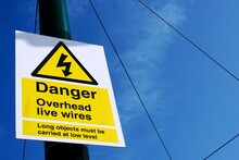 UK Sign Danger. Overhead Live Wires Against A Blue Cloudy Sky.