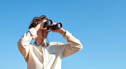 A young caucasian male looking at a view with binoculars under the bright blue sky