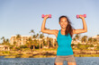 © Ariwasabi - Happy fit Asian woman training shoulders with shoulder press standing lifting free dumbbells weights at outdoor summer beach resort.