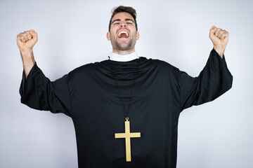 Wall Mural - Young hispanic man wearing priest uniform standing over white background very happy and excited making winner gesture with raised arms, smiling and screaming for success.