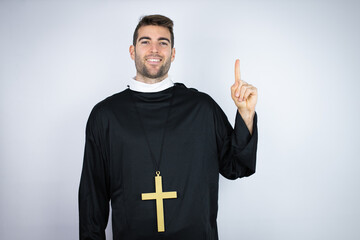 Wall Mural - Young hispanic man wearing priest uniform standing over white background showing and pointing up with fingers number one while smiling confident and happy