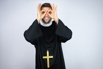 Wall Mural - Young hispanic man wearing priest uniform standing over white background doing ok gesture shocked with smiling face, eye looking through fingers