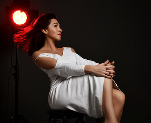 Adult pretty brunette woman actress in white dress sits on chair in neon spotlight with her hair fluttering in the wind over dark background. Fashion, stylish casual look for lady concept