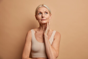 Serious tender wrinkled woman touches skin after applying anti age cream concentrated above with thoughtful expression wears cropped top isolated over brown background. Wellness hydration wellbeing