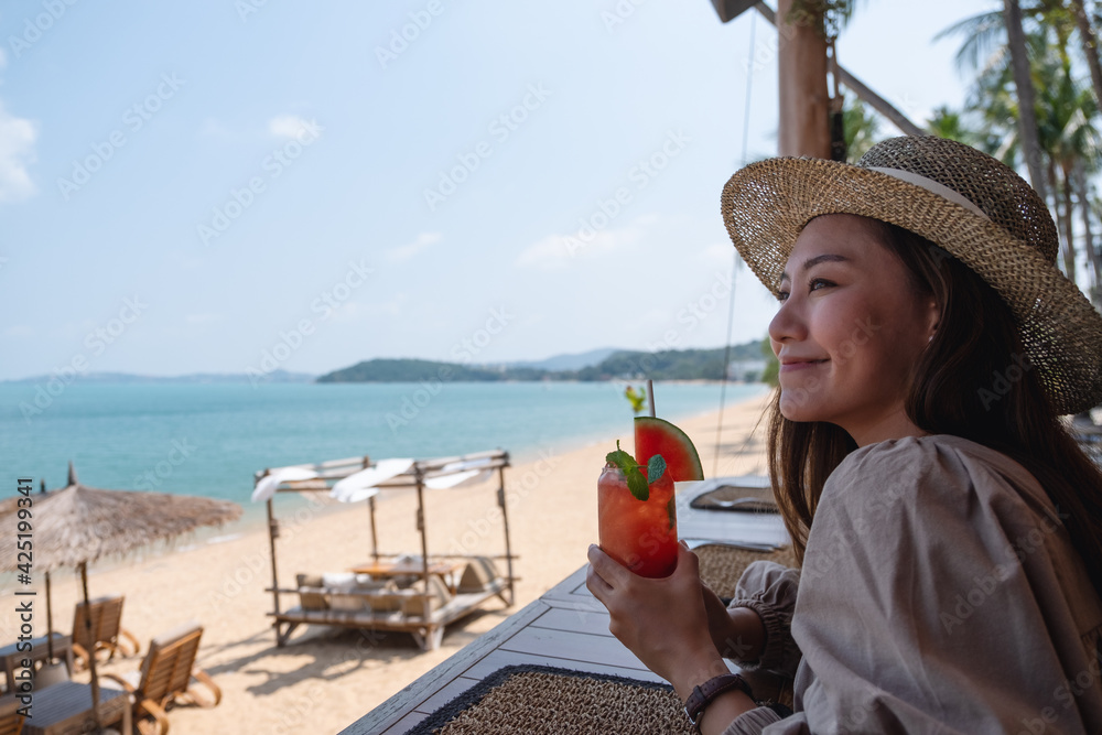 A beautiful young asian woman drinking watermelon juice while sitting in the beach cafe - obrazy, fototapety, plakaty 