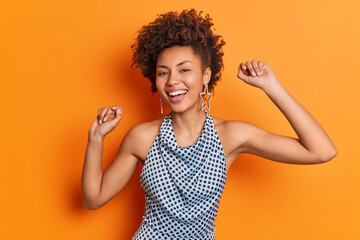 Wall Mural - Positive dark skinned curly haired lady dances with arms raised smiles broadly wears fashionable polka dot blouse isolated over vivid orange background has happy mood. People emotions joy concept