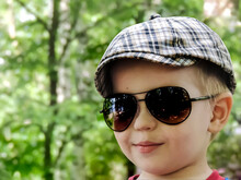 The Face Of A Smiling Little Boy In A Checkered Cap And Black Sunglasses On A Green Background.
