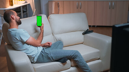 Poster - Caucasian male looking phone with at mock up green screen chroma key display while lying on sofa browsing on internet. Caucasian male using modern technology wireless late at night in kitchen