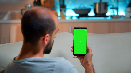 Poster - Caucasian male reading online social information on phone with mock up green screen chroma key display. Tired man sitting on sofa using isolated smartphone with technology internet