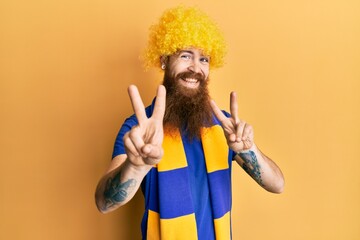 Wall Mural - Redhead man with long beard football hooligan cheering game wearing funny wig smiling looking to the camera showing fingers doing victory sign. number two.