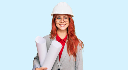 Canvas Print - Young redhead architect woman wearing safety helmet holding blueprints looking positive and happy standing and smiling with a confident smile showing teeth