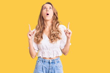 Poster - Young caucasian woman with blond hair wearing elegant summer tshirt amazed and surprised looking up and pointing with fingers and raised arms.