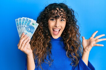 Young hispanic girl holding 50 polish zloty banknotes celebrating achievement with happy smile and winner expression with raised hand