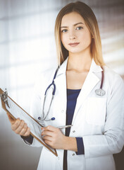 Wall Mural - Young woman-doctor is making some notes using a clipboard, while standing in her sunny cabinet in a clinic. Portrait of beautiful female physician with a stethoscope. Perfect medical service in a