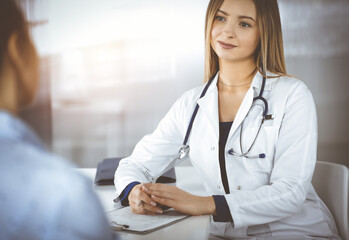 Wall Mural - Young intelligent woman-doctor is listening to her patient's allergy's symptoms, while they are sitting together in the sunny cabinet in a clinic. Female physician, with a stethoscope, is writing at