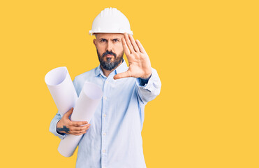 Poster - Young handsome man wearing hardhat holding paper blueprints with open hand doing stop sign with serious and confident expression, defense gesture