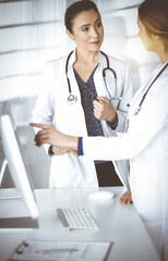 Wall Mural - Two female physicians are discussing medical therapy, while standing at the table in a sunny clinic office. Doctors use pc computer at work. Teamwork in medicine