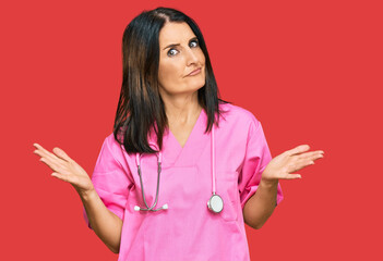 Sticker - Middle age brunette woman wearing doctor uniform and stethoscope clueless and confused expression with arms and hands raised. doubt concept.