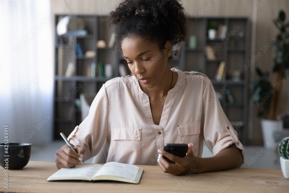 Focused young African American woman make notes handwrite in notebook use application on smartphone. Millennial biracial female write make list plan with cellphone. Planner, management concept. - obrazy, fototapety, plakaty 
