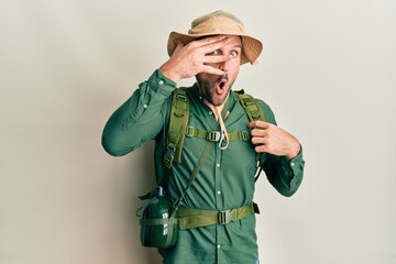 Canvas Print - Handsome man with beard wearing explorer hat and backpack peeking in shock covering face and eyes with hand, looking through fingers with embarrassed expression.