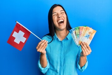 Poster - Young latin woman holding switzerland flag and franc banknotes celebrating crazy and amazed for success with open eyes screaming excited.