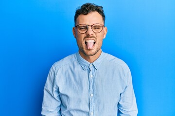 Wall Mural - Young hispanic man wearing casual clothes and glasses sticking tongue out happy with funny expression. emotion concept.