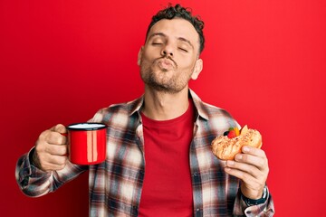 Poster - Young hispanic man drinking coffee and eating pastry looking at the camera blowing a kiss being lovely and sexy. love expression.