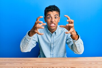 Poster - Young handsome african american man wearing casual clothes sitting on the table shouting frustrated with rage, hands trying to strangle, yelling mad