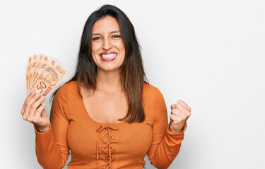 Beautiful hispanic woman holding 50 brazilian real banknotes screaming proud, celebrating victory and success very excited with raised arms