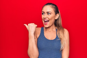 Sticker - Young beautiful blonde woman wearing sporty t-shirt standing over isolated red background pointing thumb up to the side smiling happy with open mouth