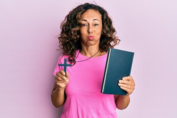 Sticker - Middle age hispanic woman holding bible and christian cross puffing cheeks with funny face. mouth inflated with air, catching air.