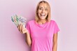 © Krakenimages.com - Beautiful blonde woman holding singapore dollars banknotes looking positive and happy standing and smiling with a confident smile showing teeth
