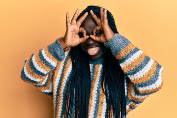 Poster - Young black woman with braids wearing casual winter sweater doing ok gesture like binoculars sticking tongue out, eyes looking through fingers. crazy expression.