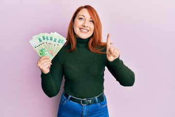 Wall Mural - Beautiful redhead woman holding 50 hong kong dollars banknotes smiling with an idea or question pointing finger with happy face, number one