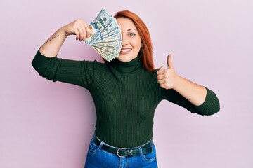 Poster - Beautiful redhead woman holding dollars banknote over eye smiling happy and positive, thumb up doing excellent and approval sign