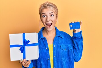 Poster - Young blonde girl holding gift and credit card celebrating crazy and amazed for success with open eyes screaming excited.