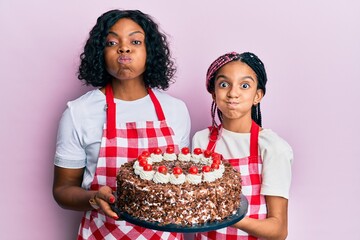 Poster - Beautiful african american mother and daughter wearing baker apron holding homemade cake puffing cheeks with funny face. mouth inflated with air, catching air.